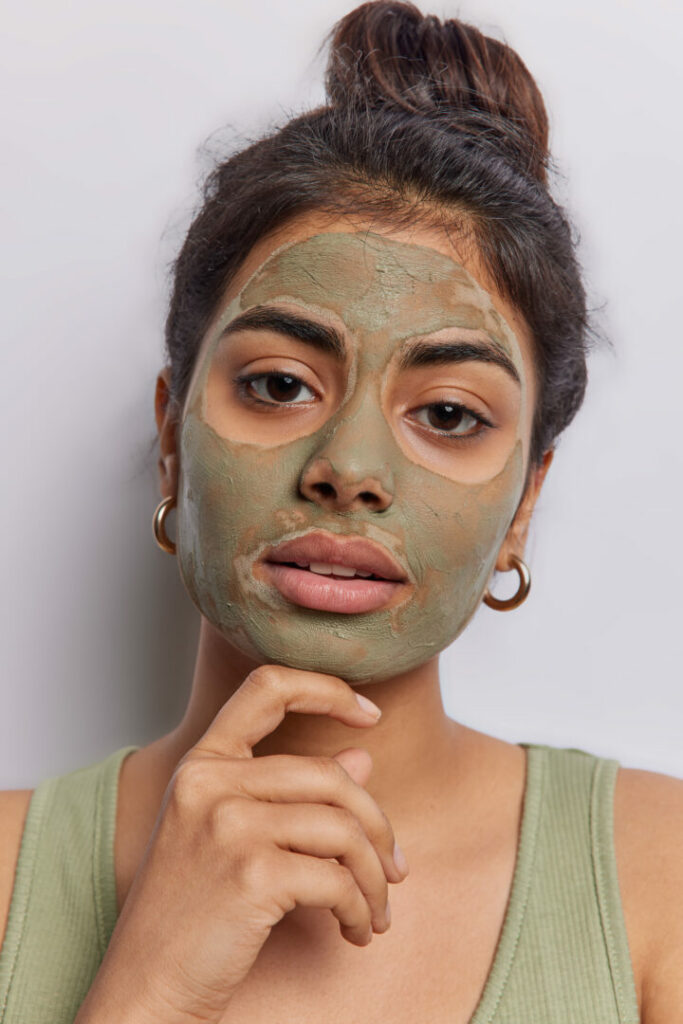 Vertical shot of serious Indian woman applies anti aging moisturising face mask for healthy skin touches chin gently focused directly at camera wears t shirt isolated on white wall. Facial treatments